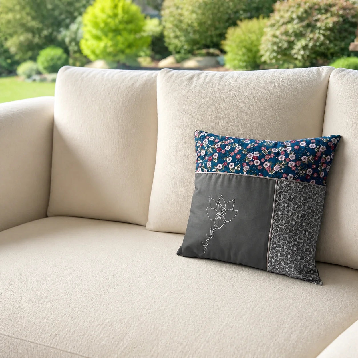 Coussin Fleur de Cerisier brodé posé sur un canapé écru devant un jardin flouté