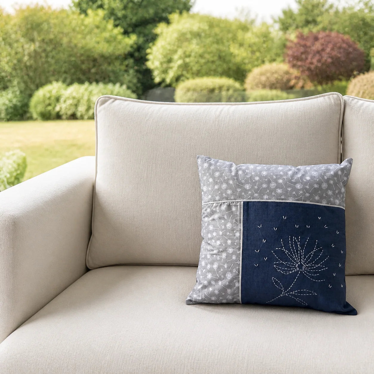 Coussin Fleur de Printemps brodé posé sur un canapé écru devant un jardin flouté