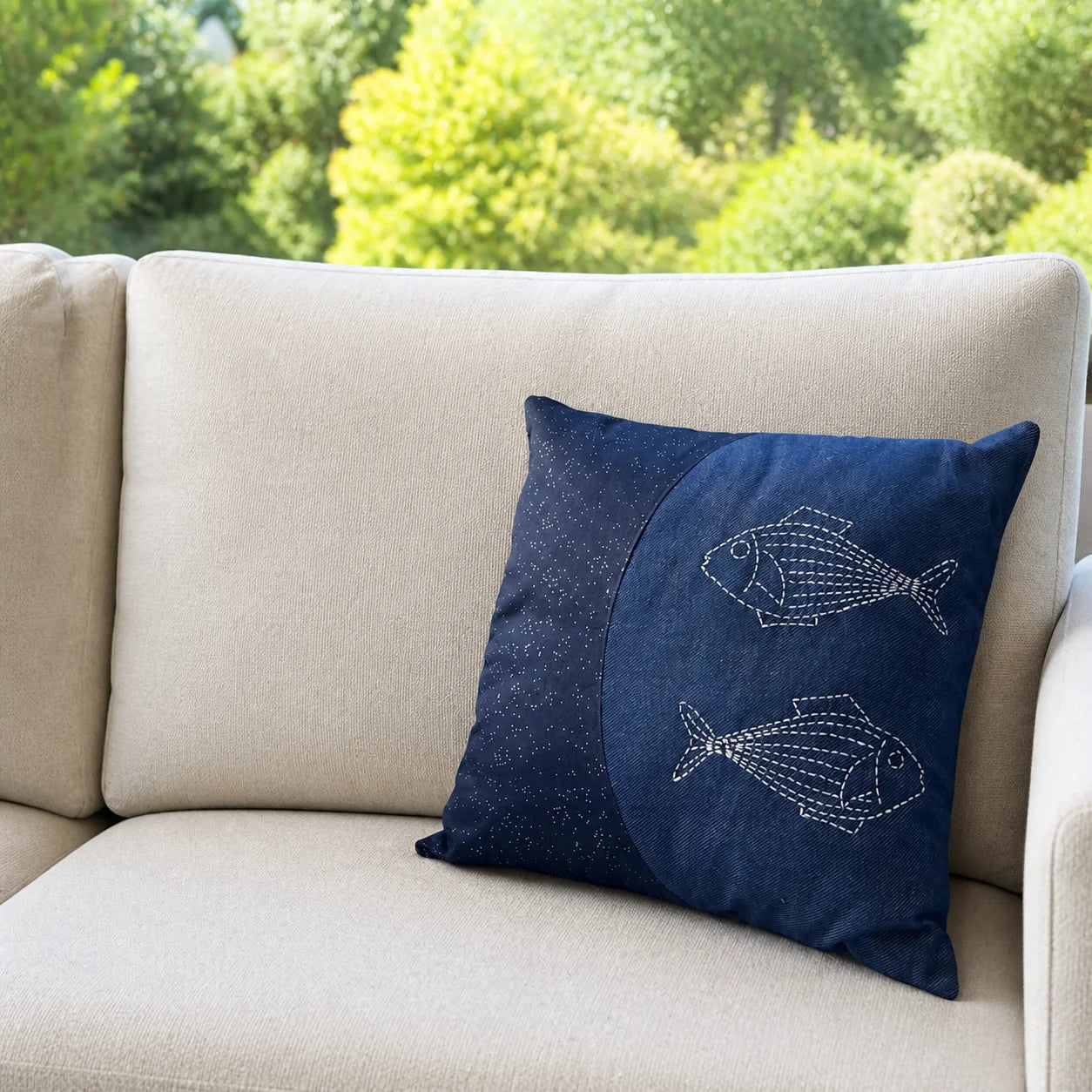 Coussin Les Deux Poissons brodé posé sur un canapé écru devant un jardin flouté