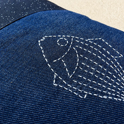 Détail d’un poisson brodé au sashiko blanc sur le denim bleu du coussin Les Deux Poissons