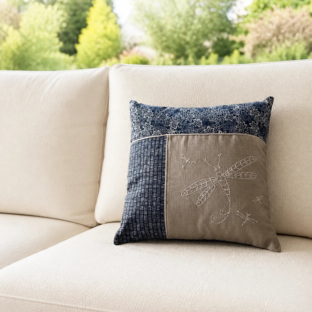 Coussin Libellule brodé posé sur un canapé écru devant un jardin flouté
